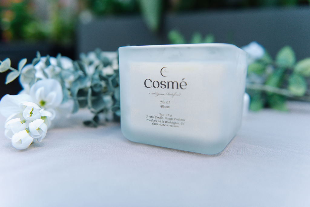 A hand-poured scented candle in the scent of Bloom 16oz - 453g is burning indoors, filling the room with a floral aroma.