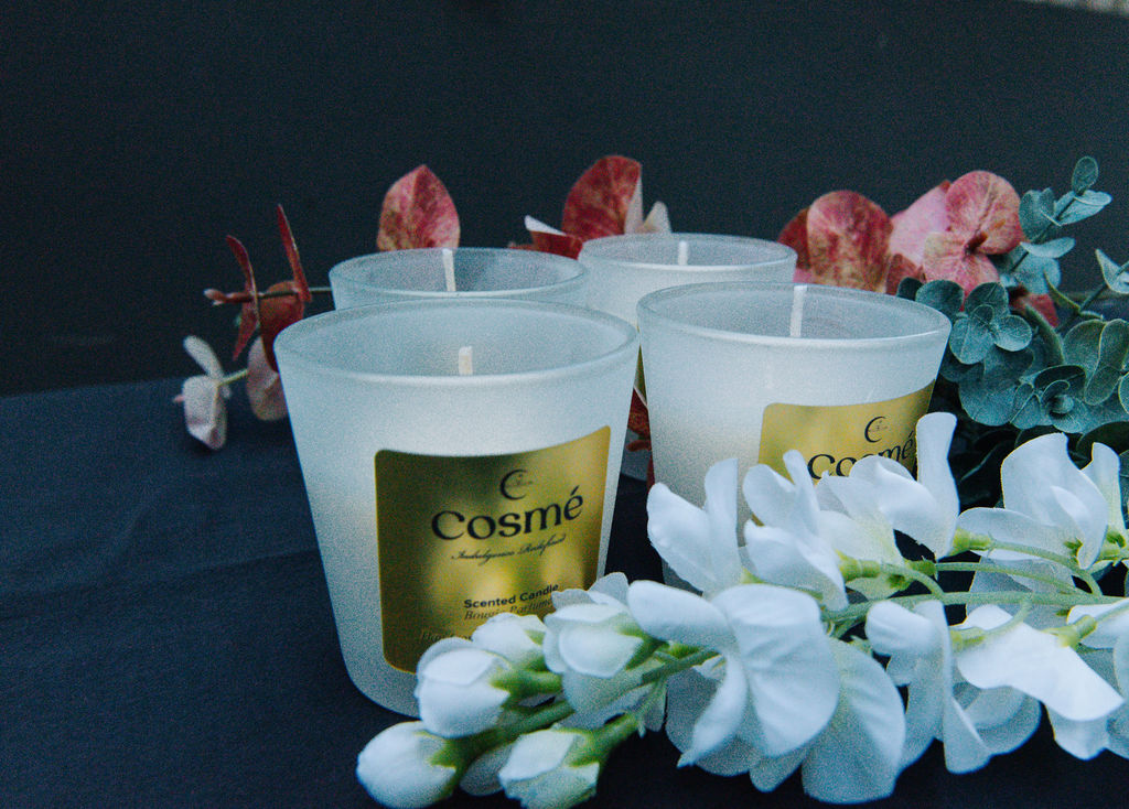 A rose-scented candle sits atop a table with two cups of drinkware, surrounded by a bouquet of fresh flowers.